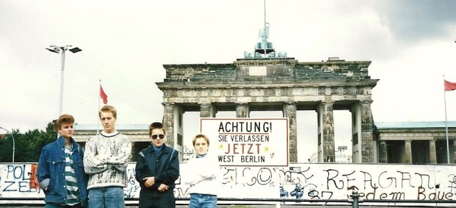 four teenagers stand in front of the Berlin Wall, with the Brandenburg Gate visible behind it