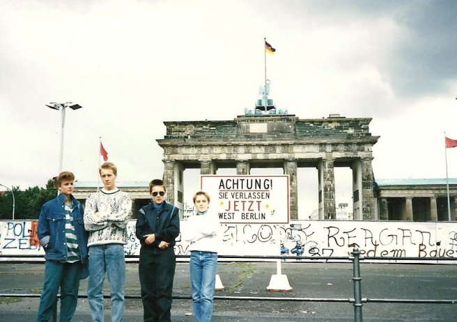 four teenagers stand in front of the Berlin Wall, with the Brandenburg Gate visible behind it